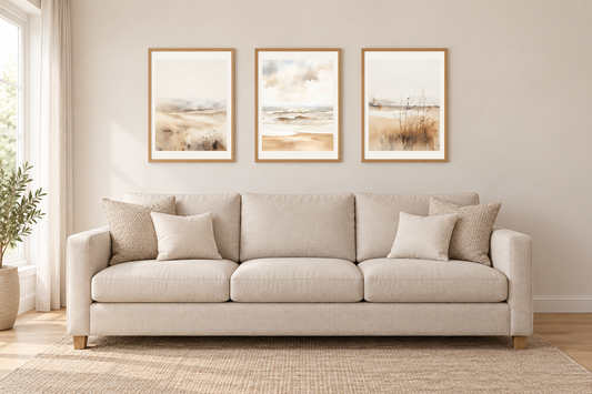 Three pale coastal prints in oak frames centered directly above a beige three-seat sofa, shown front on.