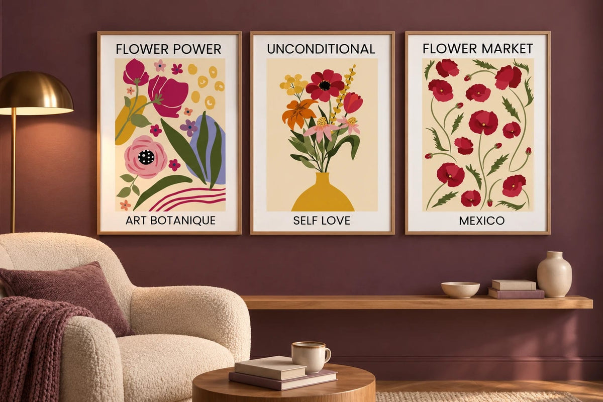 Three floral prints hang on a plum brown wall above a wood shelf beside a cream chair.
