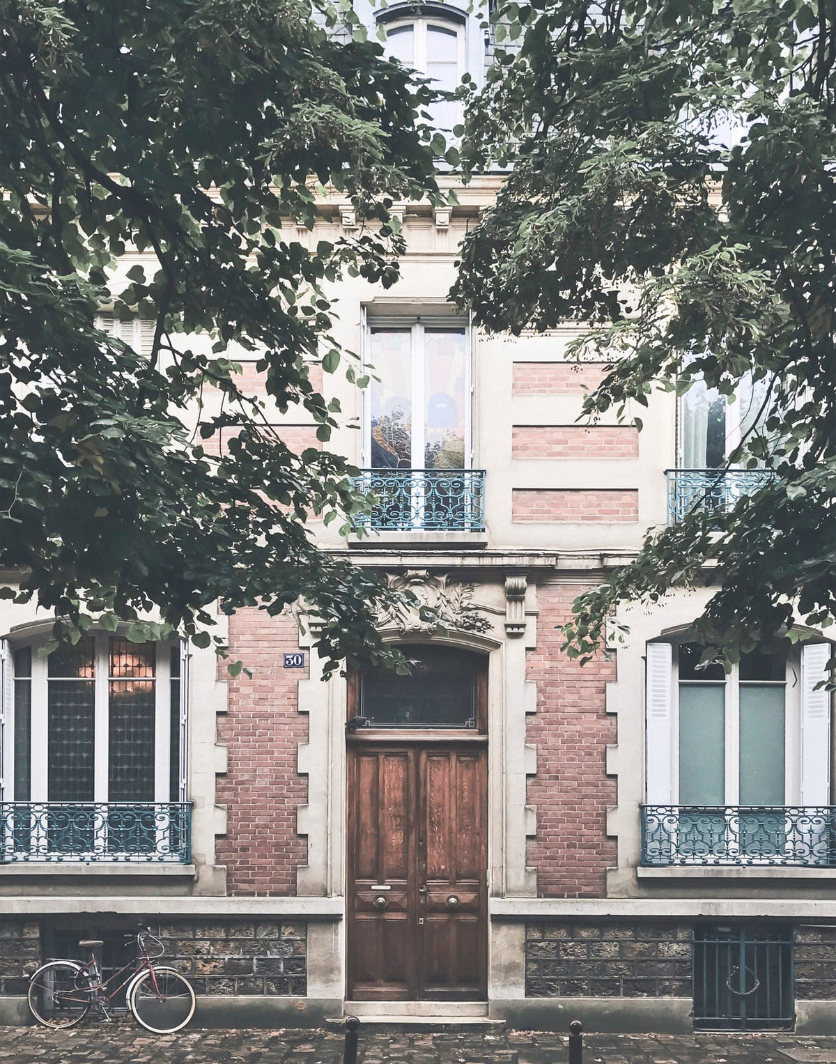 Paris doorway with faded brick, blue iron balconies, leafy branches, and a bicycle