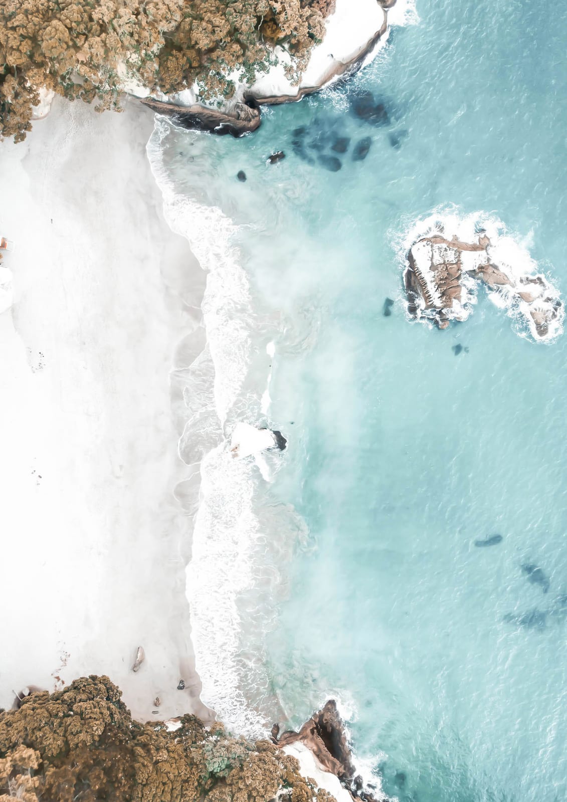 An aerial view divides pale sand from transparent turquoise water along a quiet tropical beach