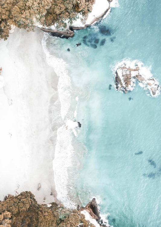 An aerial view divides pale sand from transparent turquoise water along a quiet tropical beach