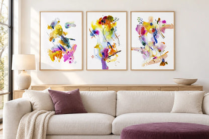 Bright abstract prints hang above a wood cabinet and light sofa against white.