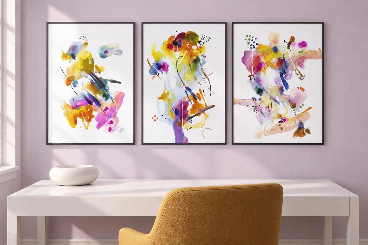Bright prints hang above a white desk with a mustard chair on a pale lavender wall.