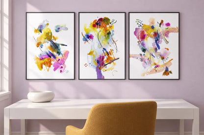 Bright prints hang above a white desk with a mustard chair on a pale lavender wall.