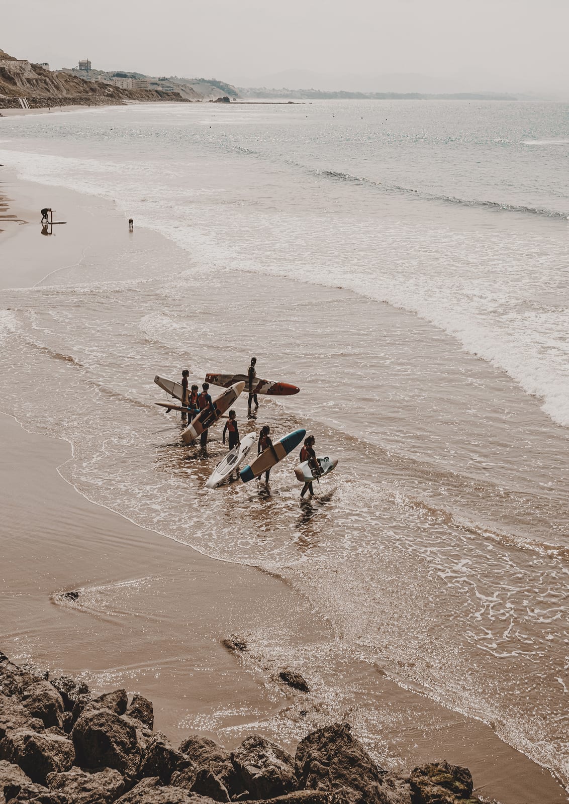 surfers carrying boards through shallow water by dark rocks.