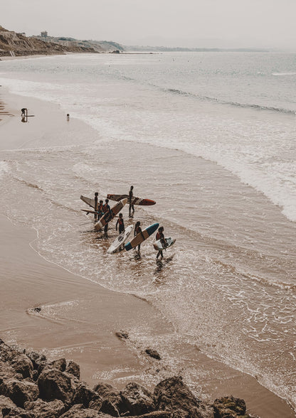 surfers carrying boards through shallow water by dark rocks.