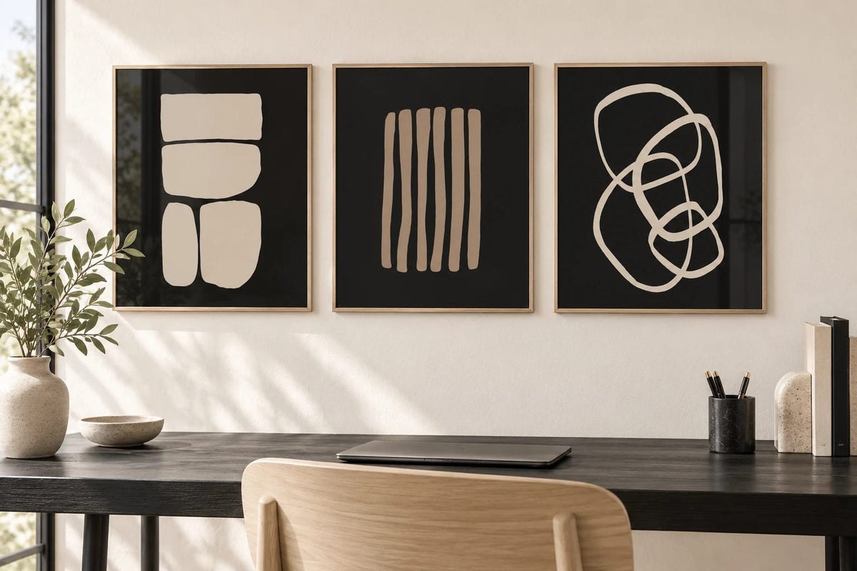 Three prints above a dark writing desk in a light office, with a laptop and books.