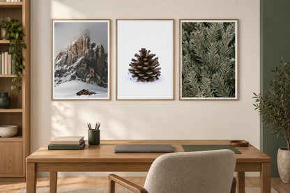 A snowy cabin, pinecone, and evergreen prints hang above an oak table in a home office.