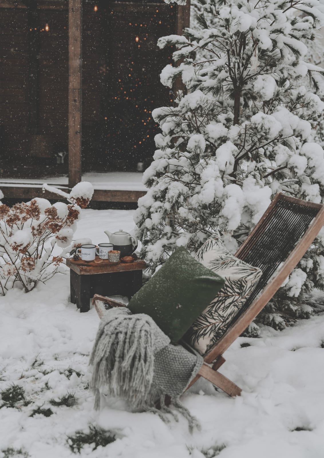 An outdoor chair with a blanket sits by a table and snowy fir.