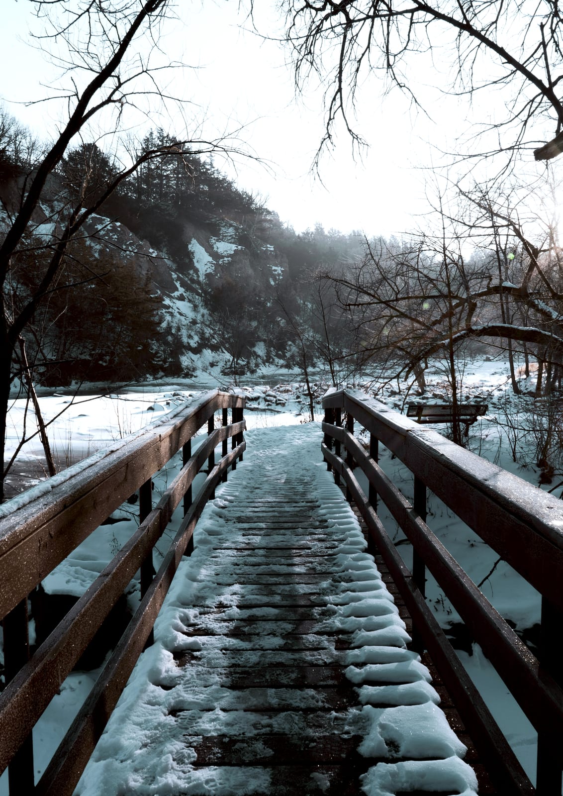 A snow-covered wooden bridge leads toward a forest scene.
