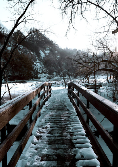 A snow-covered wooden bridge leads toward a forest scene.