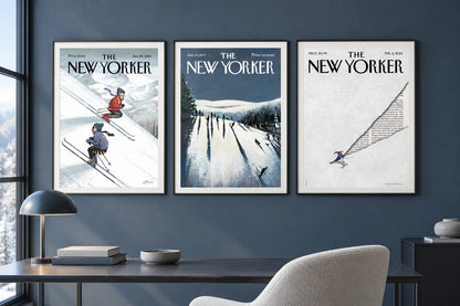 Three ski prints in dark frames hang on a blue office wall beside a dark desk and light chair.