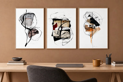 Three abstract prints hang above a wood desk on a terracotta wall.