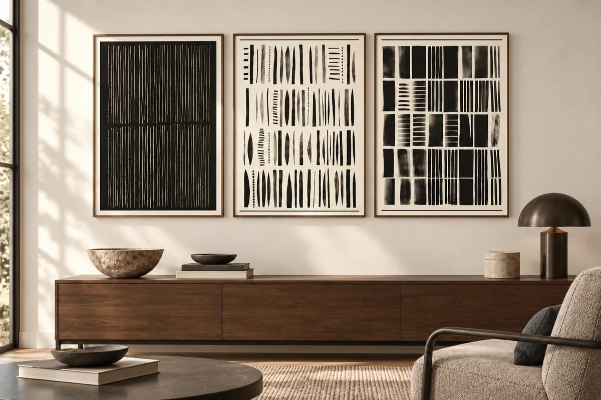 Three geometric prints hang above a light wood cabinet beside a cream chair.